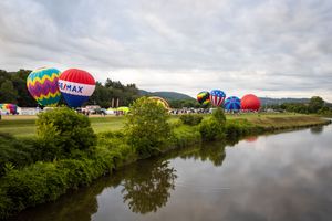 Colorful hot air balloons reflected in calm water with trees along shoreline.