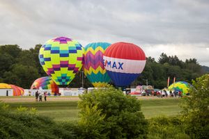 Multiple hot air balloons in vibrant patterns prepare for takeoff along a grassy field under cloudy skies.