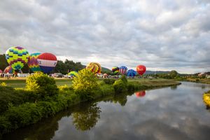 Colorful hot air balloons float above a scenic river landscape during a balloon festival on a cloudy day.