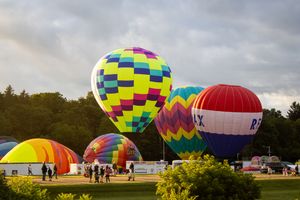 Hot air balloons in checkered and geometric patterns line up on a field during a festival gathering.