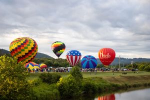 Hot air balloons in patriotic and geometric patterns prepare for launch beside a tranquil river.