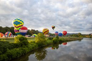 Panoramic view of hot air balloons gathering near a river with their colorful reflections in the water.