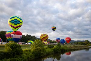 Colorful hot air balloons line the riverbank during a festival under dramatic cloudy skies.