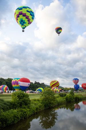Hot air balloons in bright patterns gather near a reflective river under partly cloudy skies.