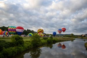Panoramic view of hot air balloons gathering near a reflective lake under cloudy skies.