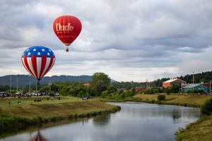 Red and white hot air balloons floating above a winding river on a cloudy day.