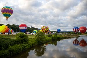 Panoramic view of hot air balloons gathering at a riverside festival under cloudy skies.