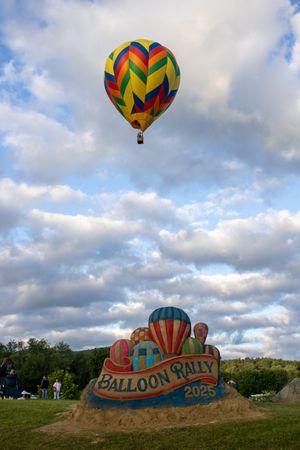 Hot air balloons with vibrant geometric patterns soaring above a sand sculpture display.