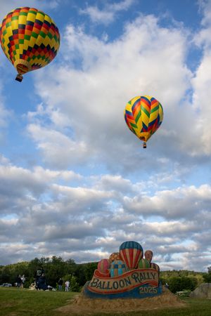 Brightly colored hot air balloons ascending over a festival grounds with sand art display.
