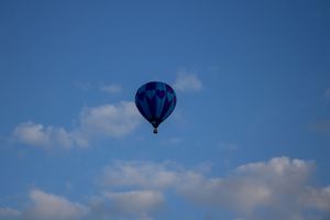 A blue hot air balloon silhouetted against bright blue sky and white clouds.