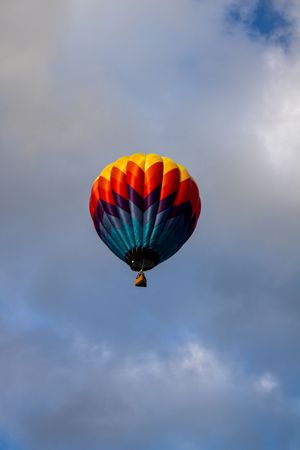 Colorful hot air balloon with red and teal panels soars through cloudy skies.