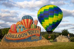 Hot air balloons take off near a decorative sand sculpture promoting a balloon festival.