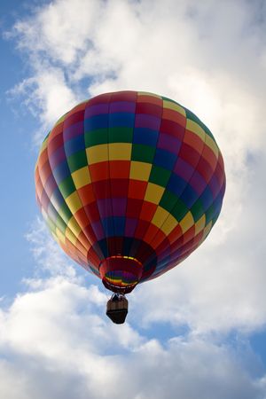 Rainbow-colored hot air balloon with geometric pattern flies through cloudy sky.
