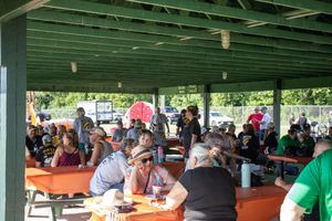 People seated at long tables under a green-roofed outdoor shelter during a community gathering.