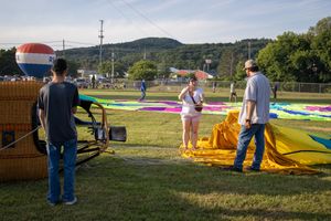Crew members prepare colorful hot air balloons for launch on a grassy field with hills in the background.