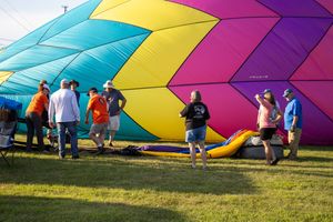 Colorful geometric patterns on a deflated hot air balloon spread across grass as people gather around.