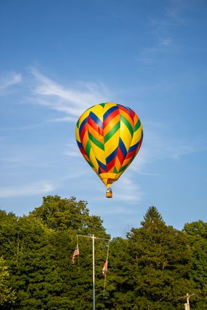 A yellow and red geometric-patterned hot air balloon floats above green trees against blue sky.
