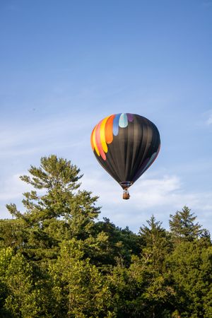 Black and orange hot air balloon floating above treeline against blue sky.