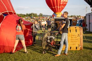 Red hot air balloons and festival crowds gathered in grassy field at sunset.