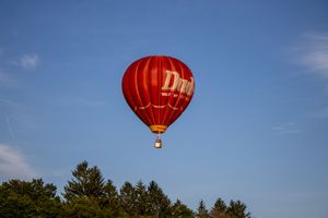 Red hot air balloon ascending into bright blue sky above treeline.