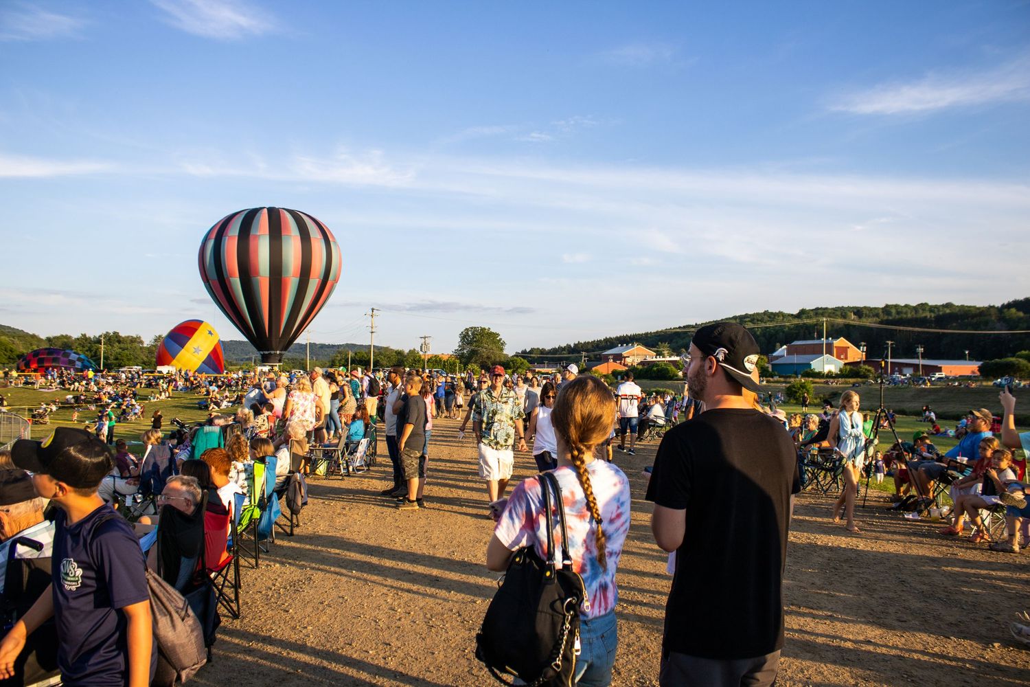 Crowd gathers at outdoor festival with hot air balloon in field under blue sky.