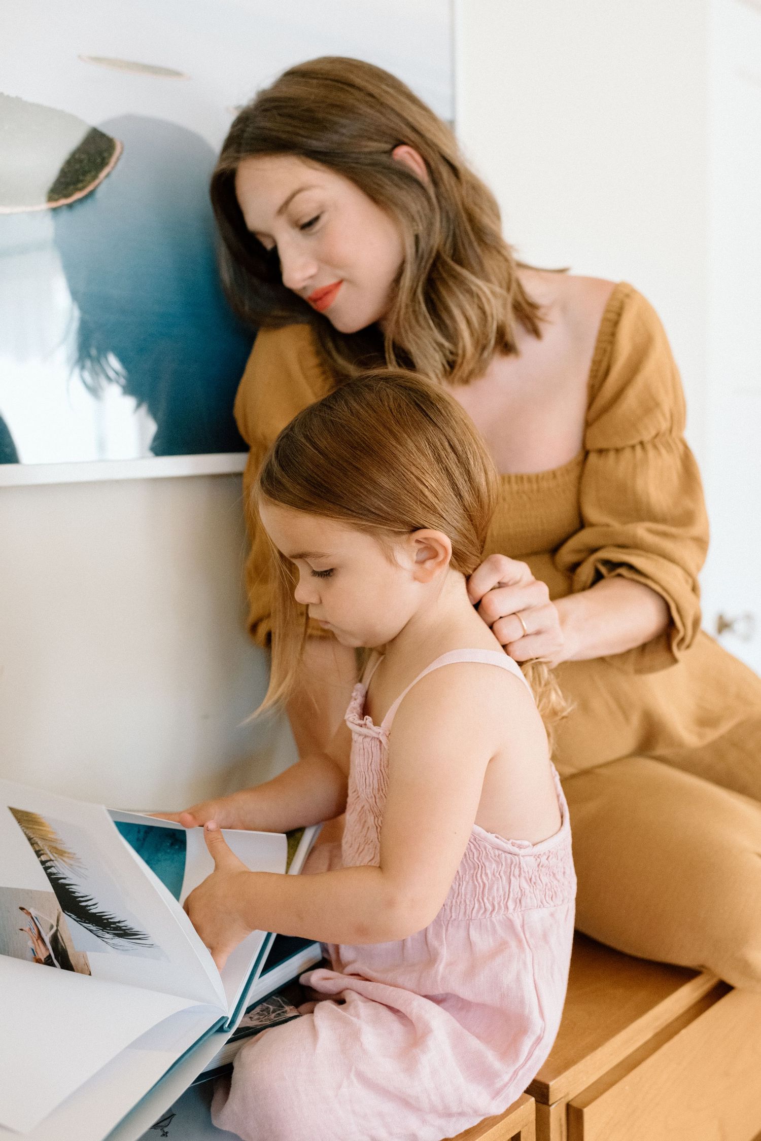 Adult and child wearing light colors read and write together at a desk in a bright interior space.
