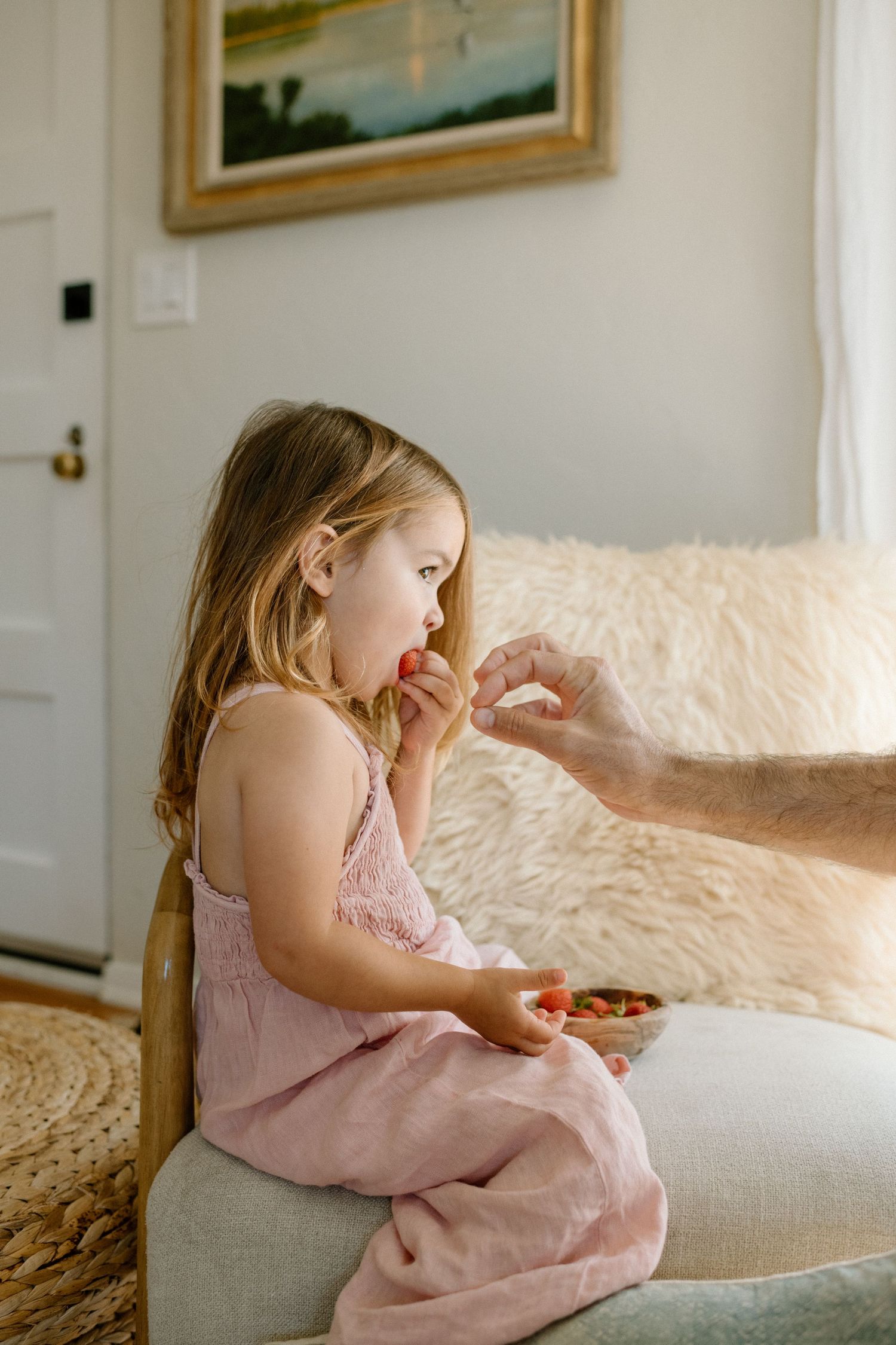 A child in a pink dress sits on a couch while a hand offers something during daytime.