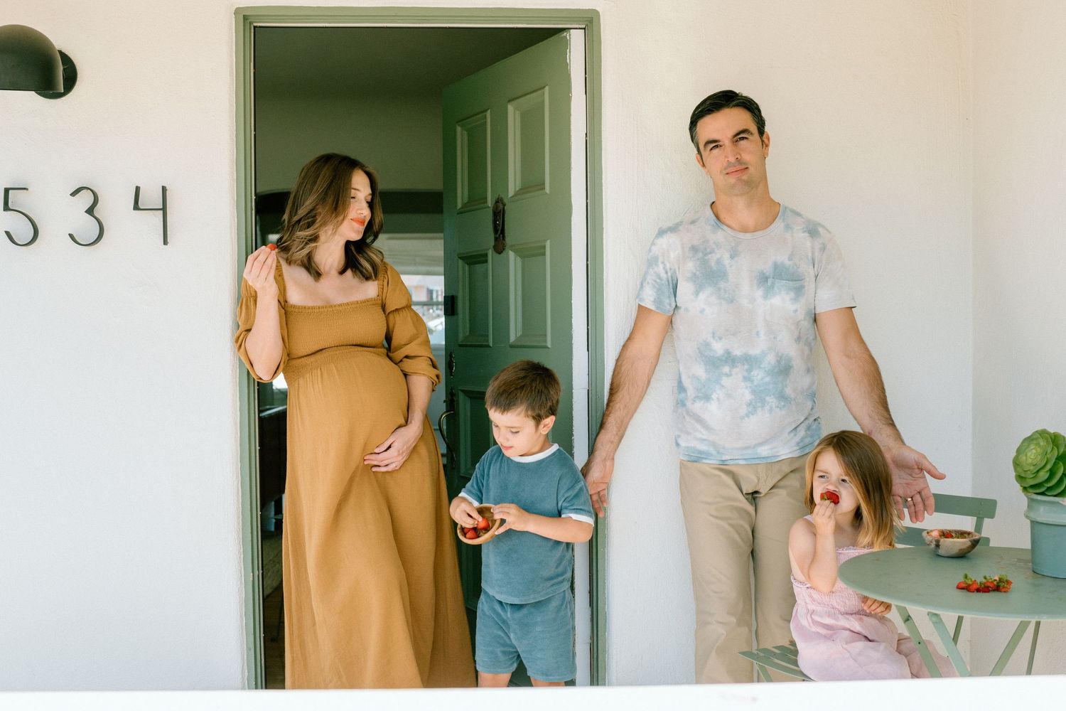 Family stands outside their front door at home address 534 with the mother wearing a mustard dress and children playing.
