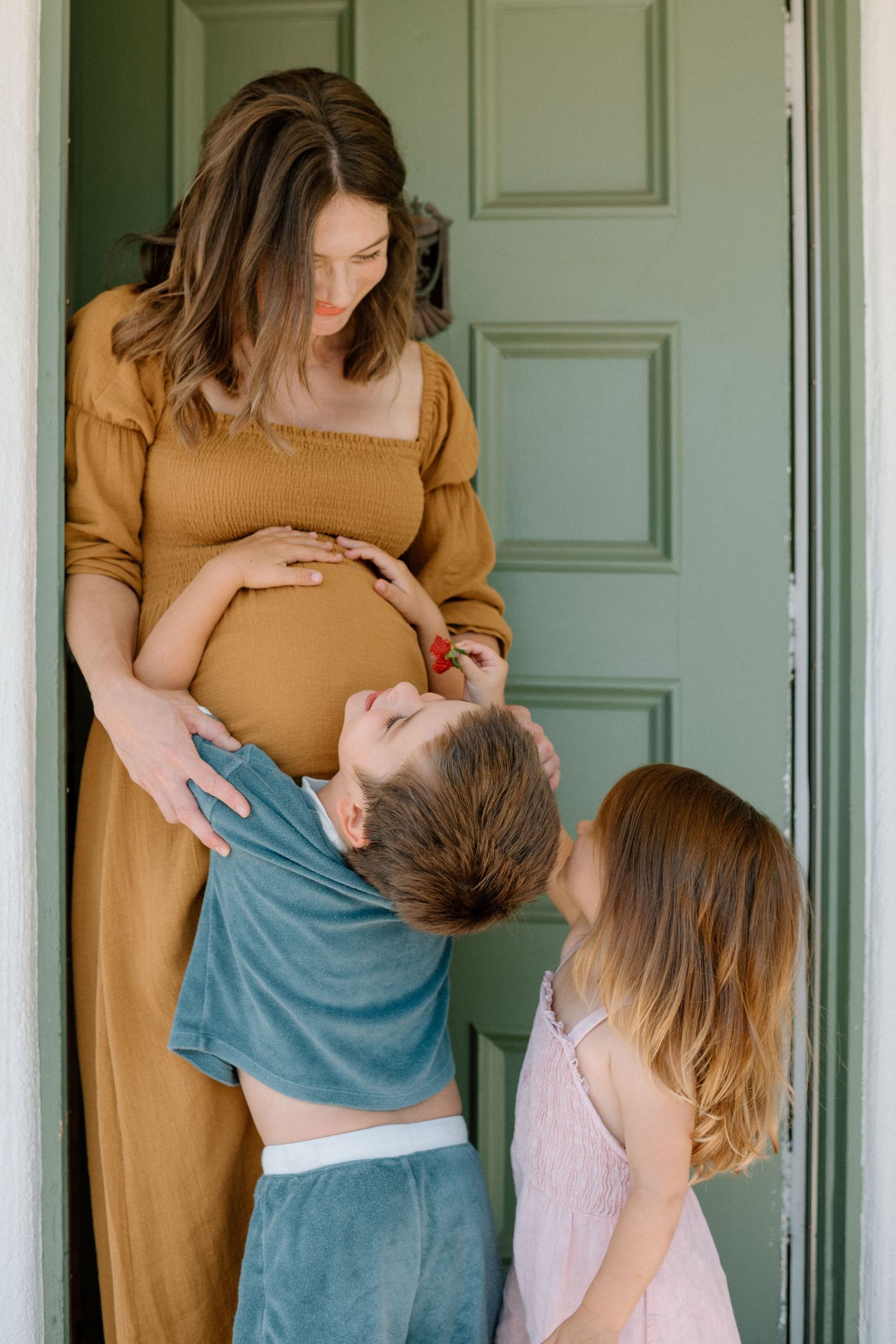 Expectant mother in mustard dress shares tender moment with child in light blue shirt against white door.