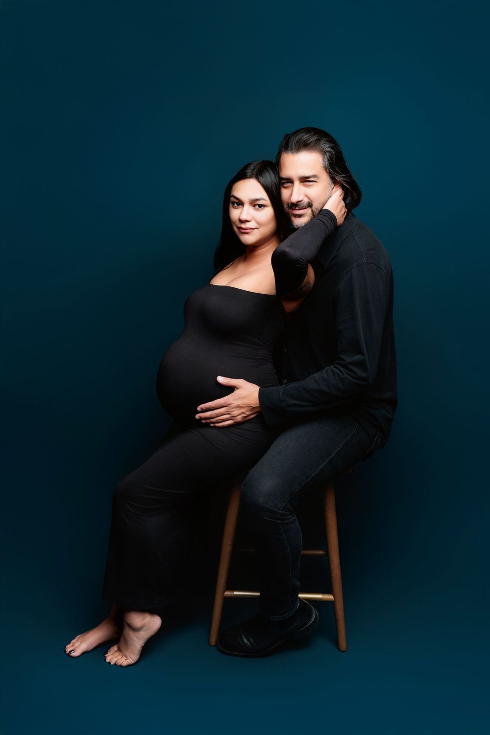 A maternity photo featuring an expectant couple in black attire against a dark turquoise background.