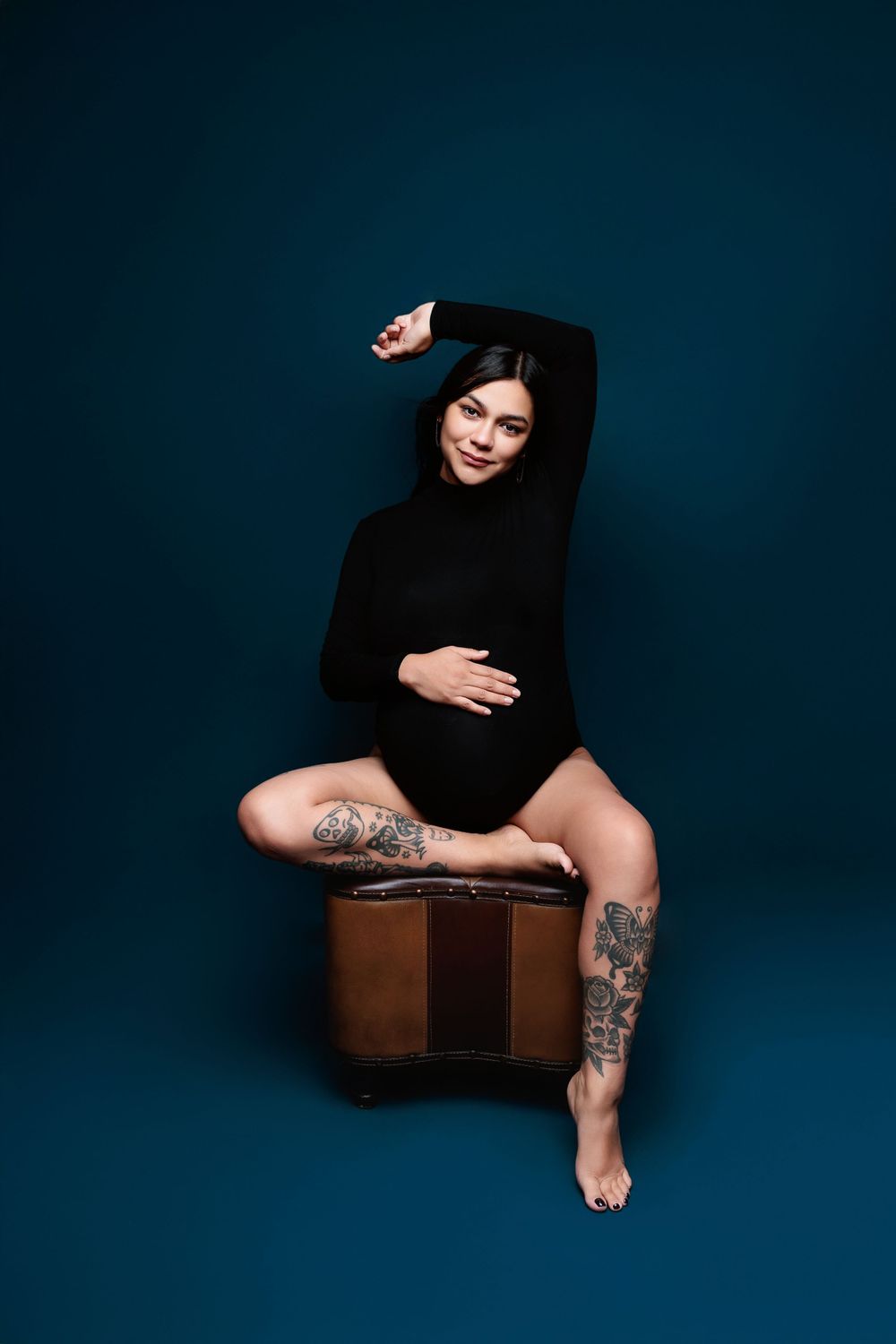 Figure in black sweater with tattoos poses artistically on wooden box against dark blue backdrop.