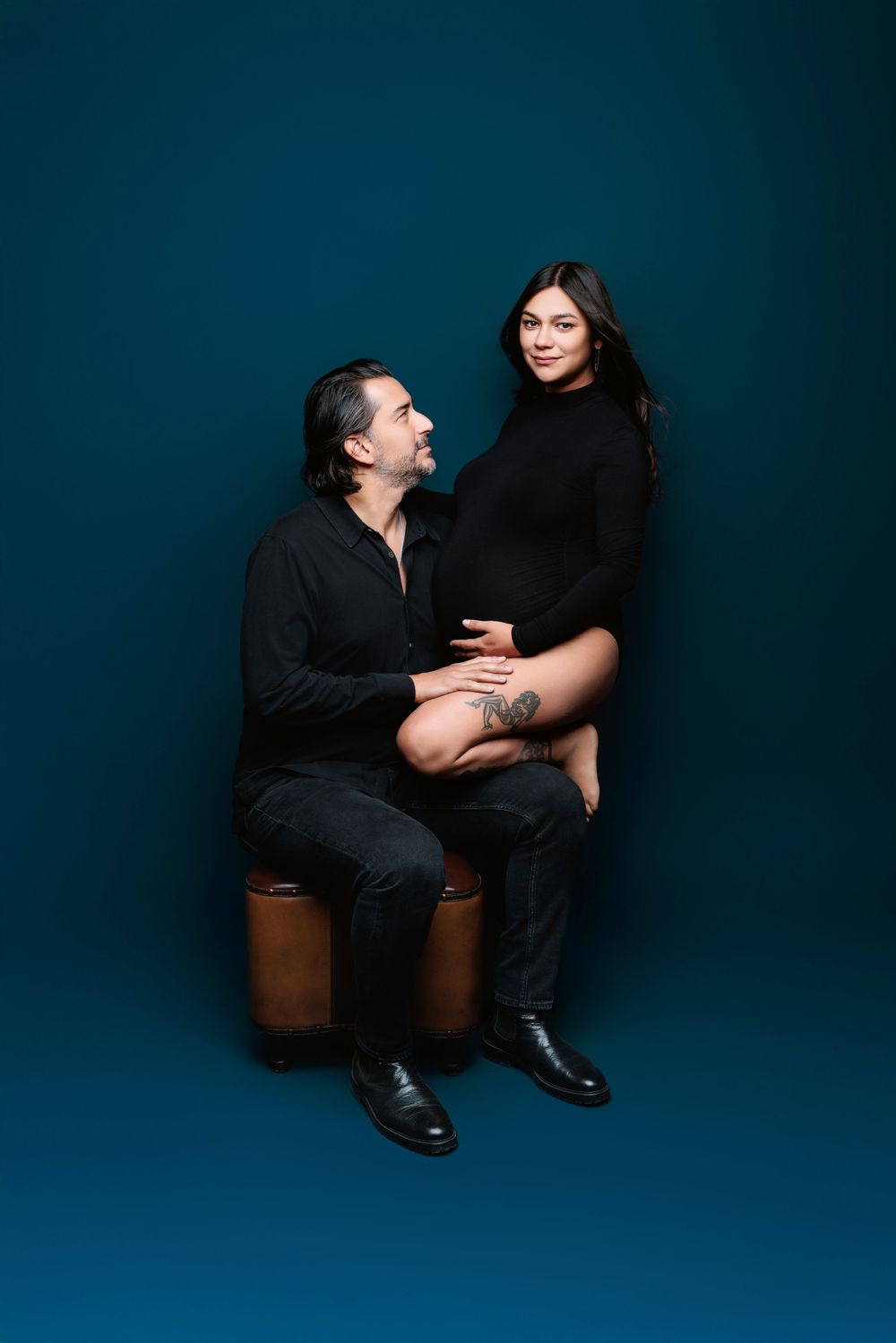 Couple in black outfits pose intimately against dark blue backdrop in studio portrait setting.