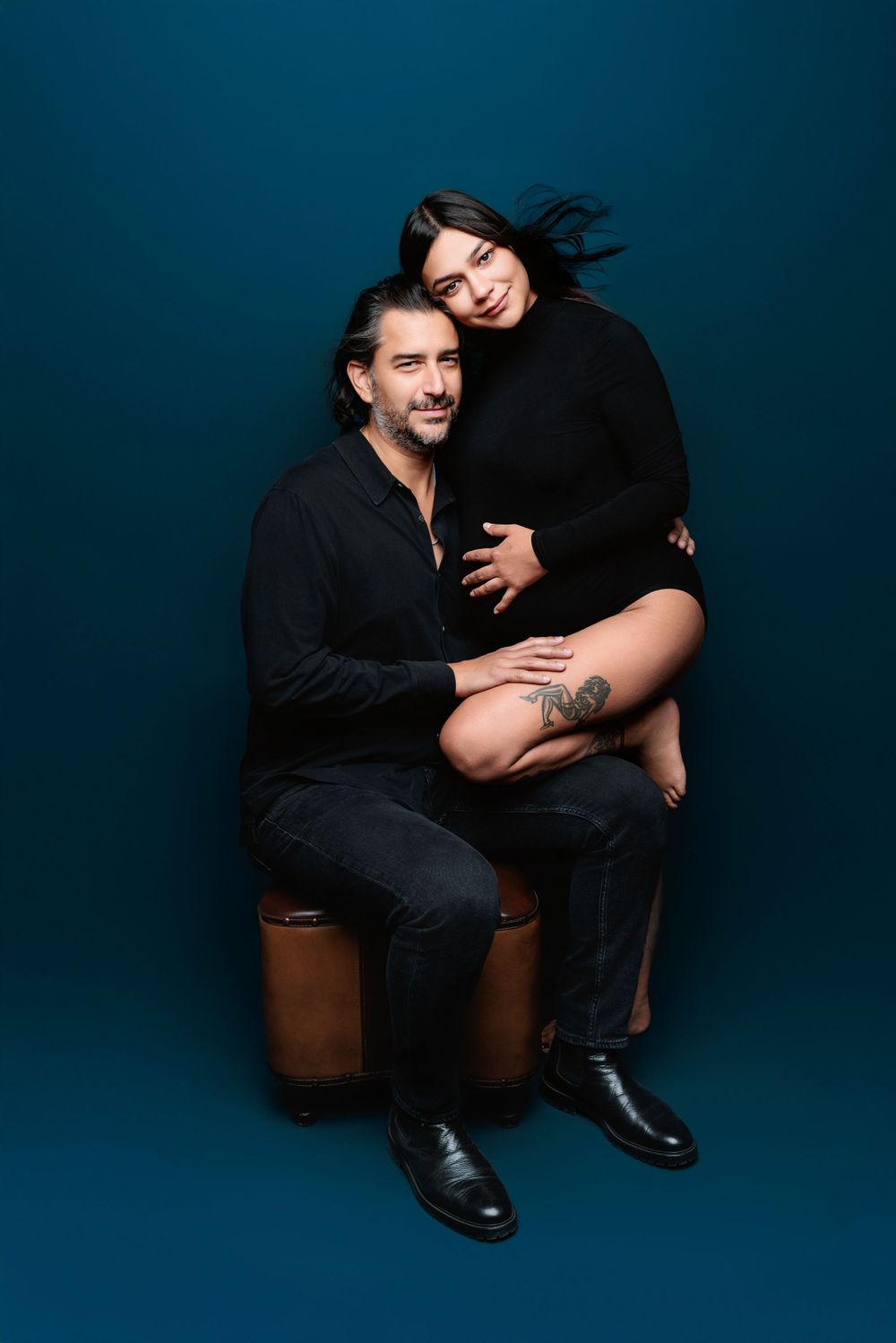 A stylish couple dressed in black poses intimately against a deep blue studio backdrop for a dramatic portrait.