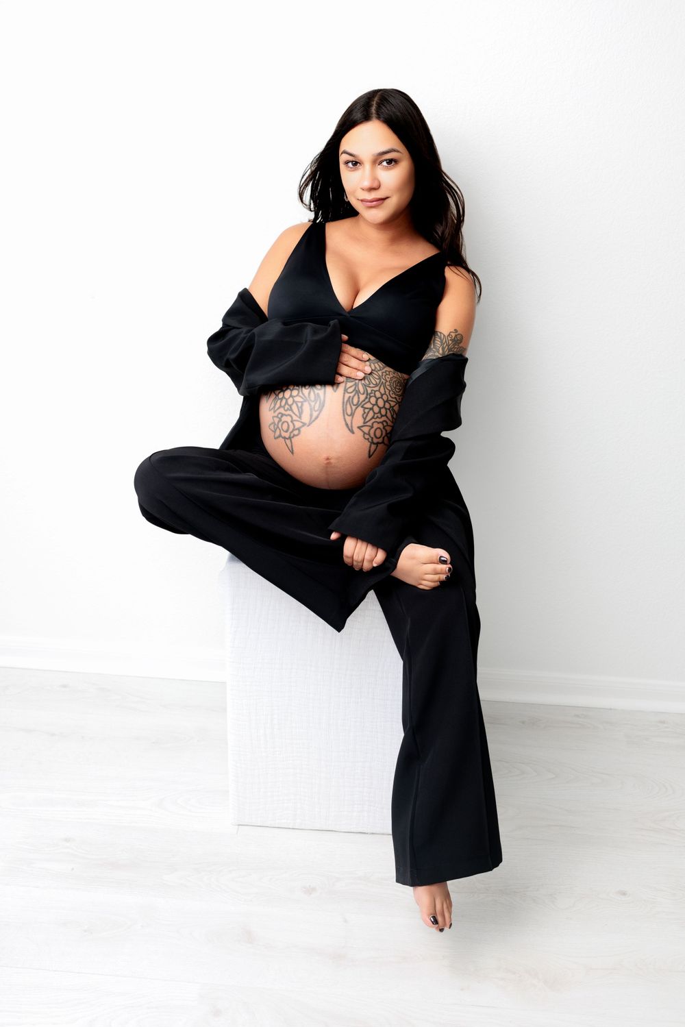 Pregnant model wearing black loungewear shows off decorated belly while posing against white wall.