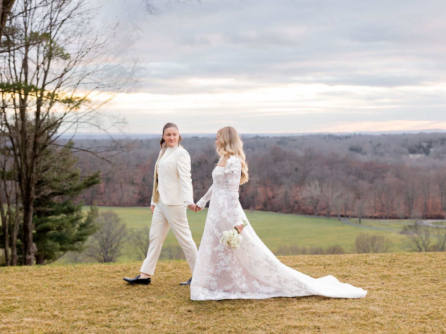 Couple in white poses on hilltop overlooking winter landscape with flowing wedding dress.