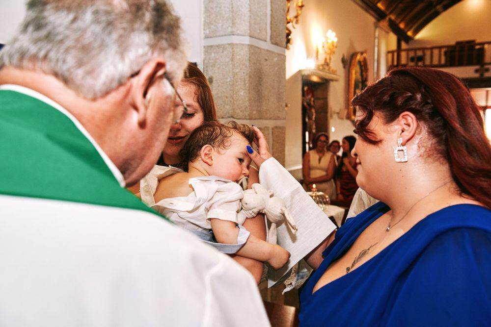 Catholic priest anointing baby with holy oil during baptism ceremony while godmother watches in blue dress.