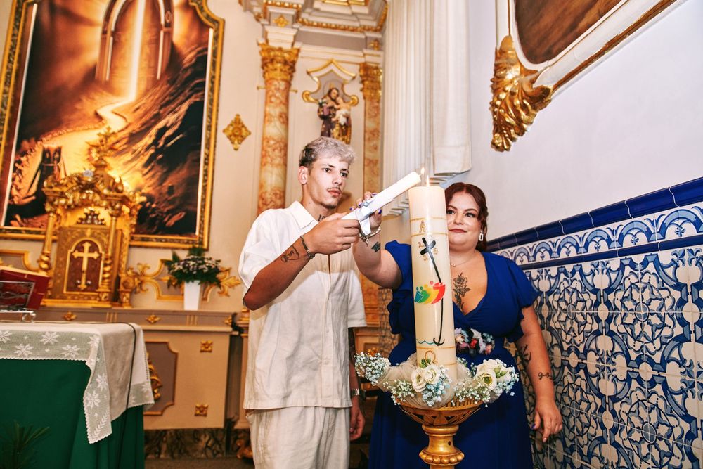 Godparents lighting a decorative baptismal candle near a floral font during a christening ceremony.