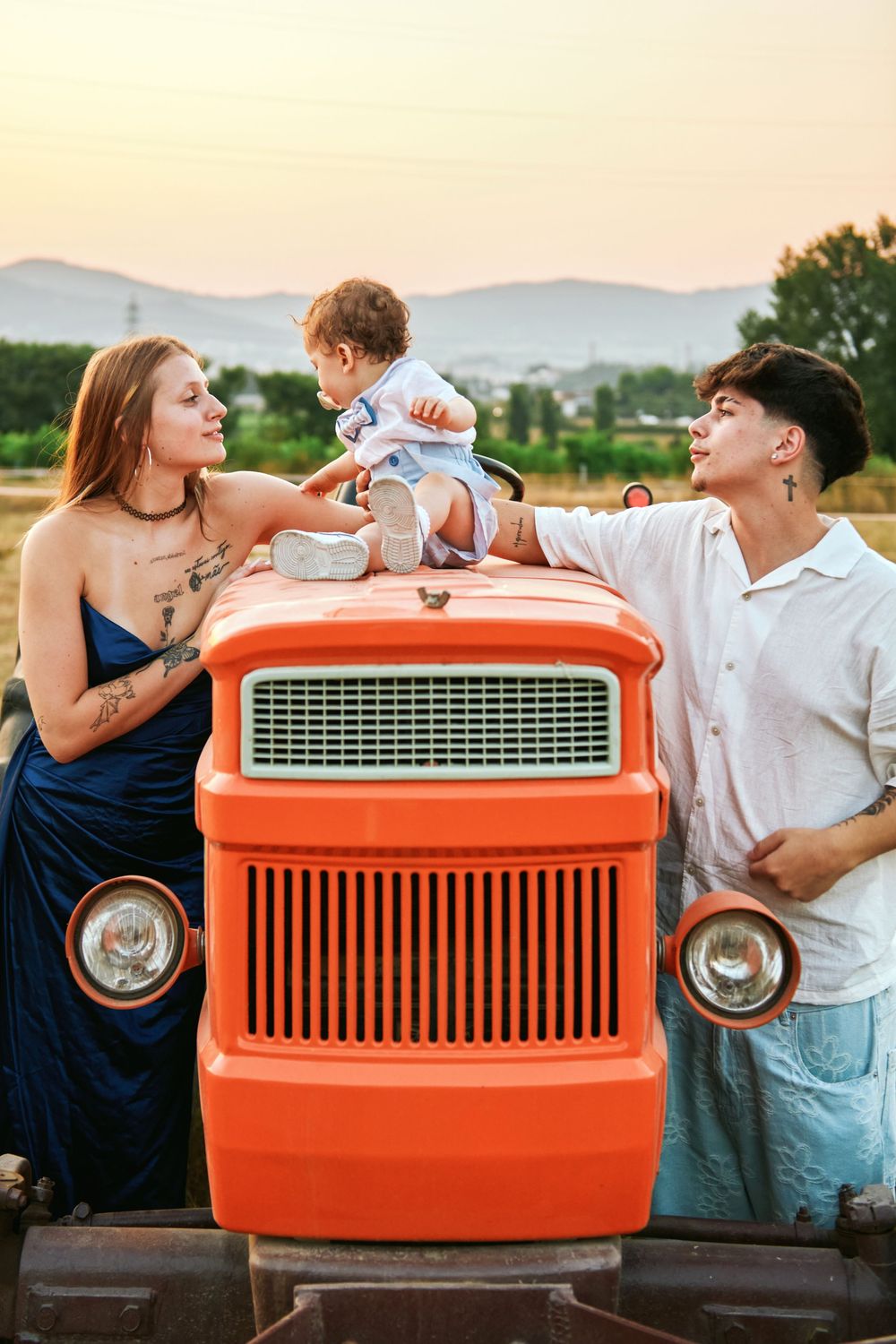 Young couple with tattoos attending to baby sitting on hood of vintage orange tractor at sunset.