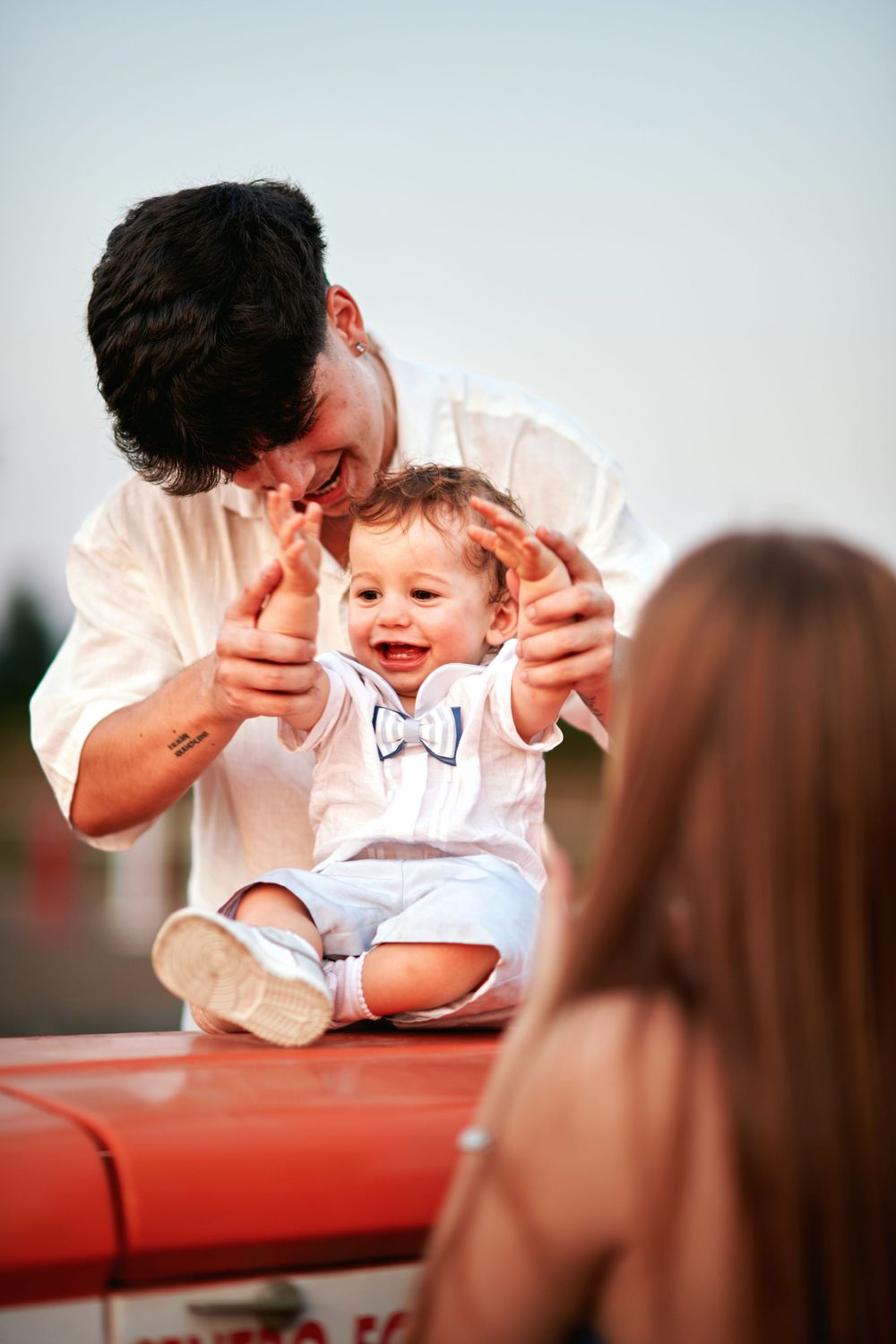 Laughing baby boy in bow tie outfit sitting on red tractor hood while father plays with him.