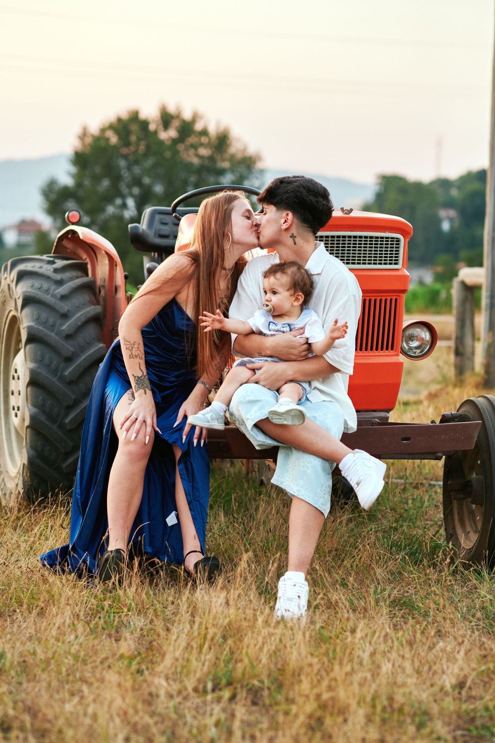 Young couple kissing while holding baby boy in front of vintage red tractor at sunset.