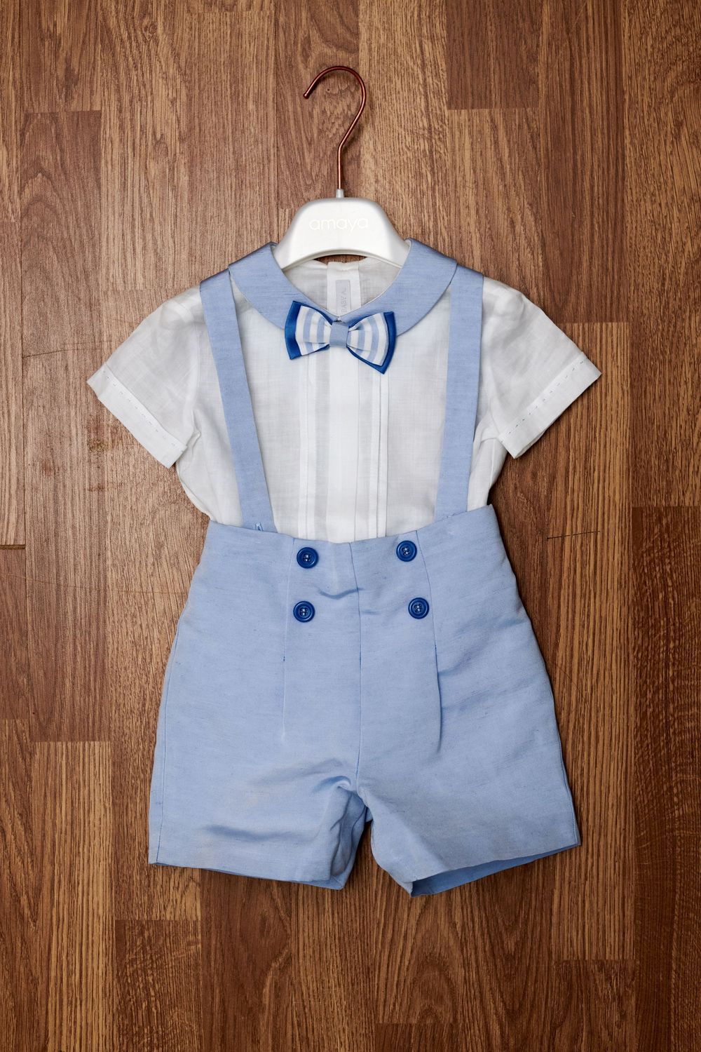 Baby boy baptism outfit with white shirt, blue suspender shorts, and blue bow tie on wooden hanger.