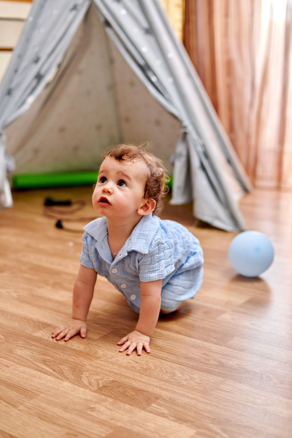 Baby boy in blue outfit crawling on hardwood floor with gray teepee tent and blue balloon in background.