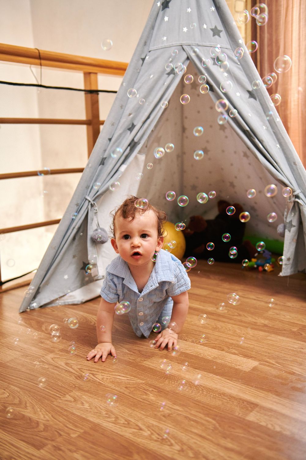 Curious baby boy crawling on wooden floor surrounded by soap bubbles near gray star teepee play tent indoors.