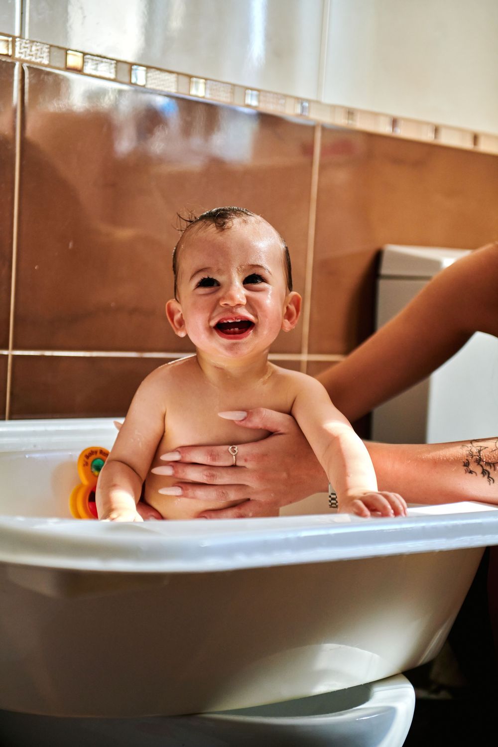 Happy smiling baby laughing during bath time in small white tub with adult hands supporting them.