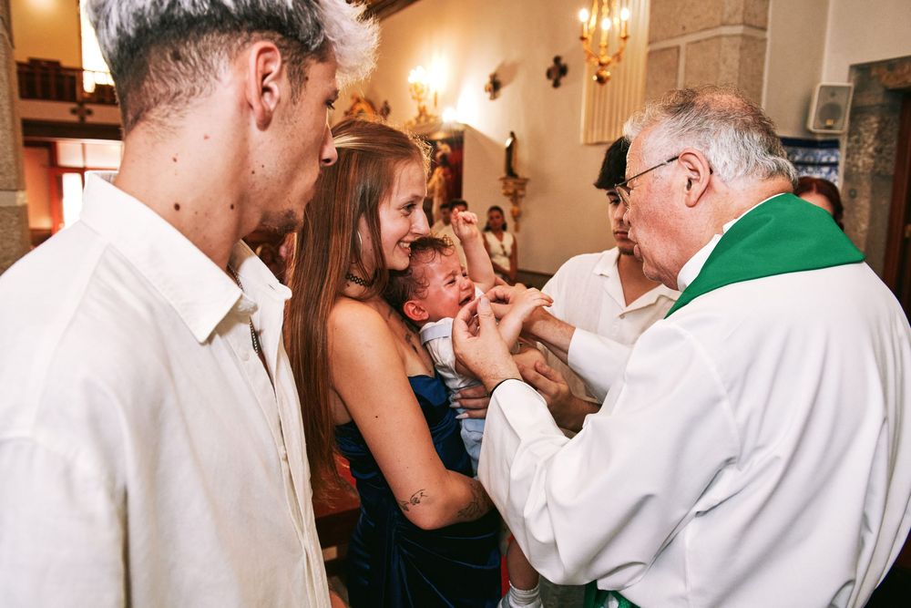 Catholic priest performing baptism rite on infant held by smiling mother inside a church ceremony.