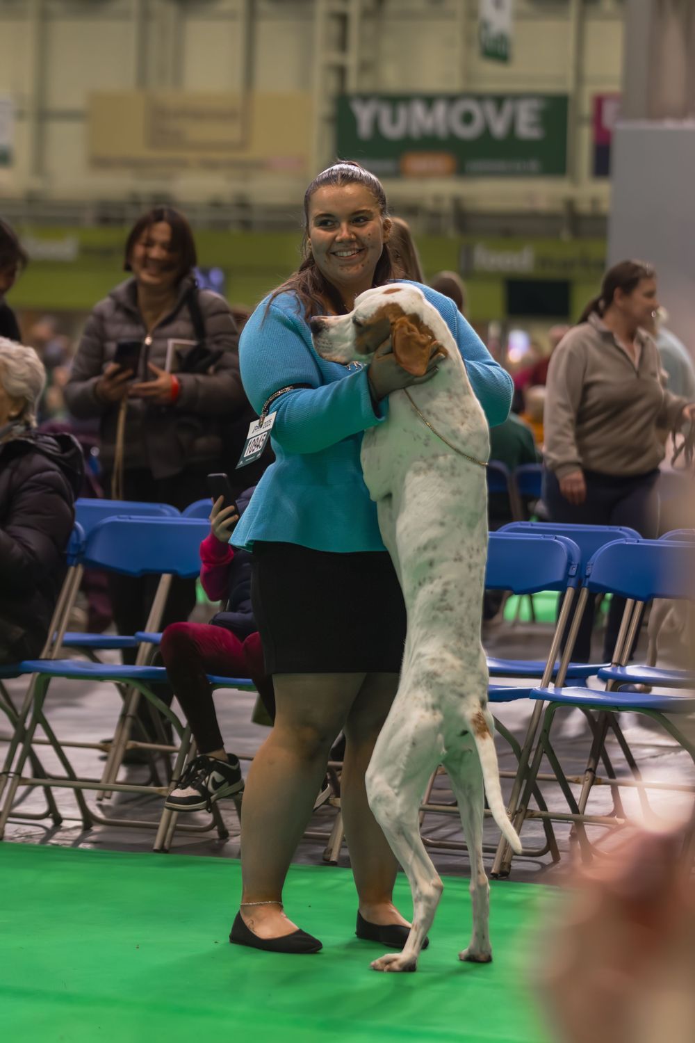 A handler in teal and black clothing dances with a large white dog on a green floor at an indoor event venue.