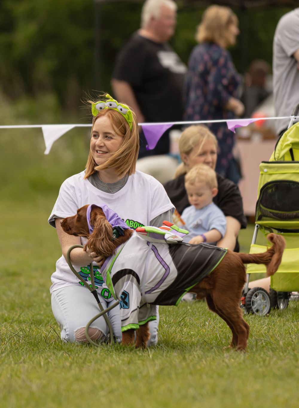 A dog wearing a colorful outfit stands with its handler on grass at an outdoor event with bunting visible in background.