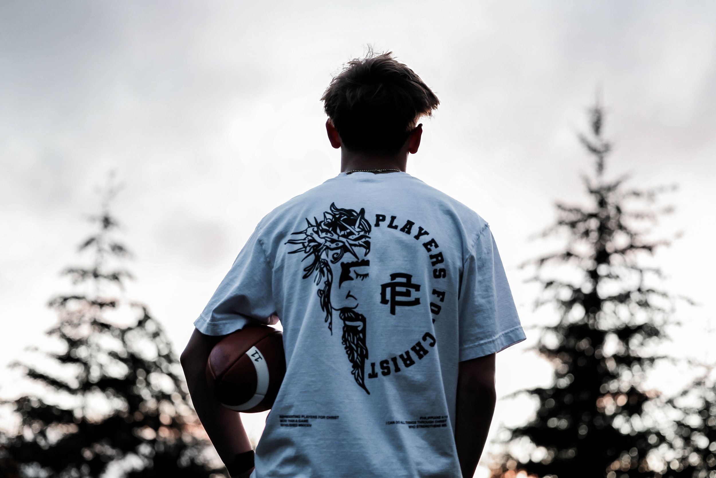 Person wearing light blue t-shirt holding football silhouetted against evergreen trees at sunset.
