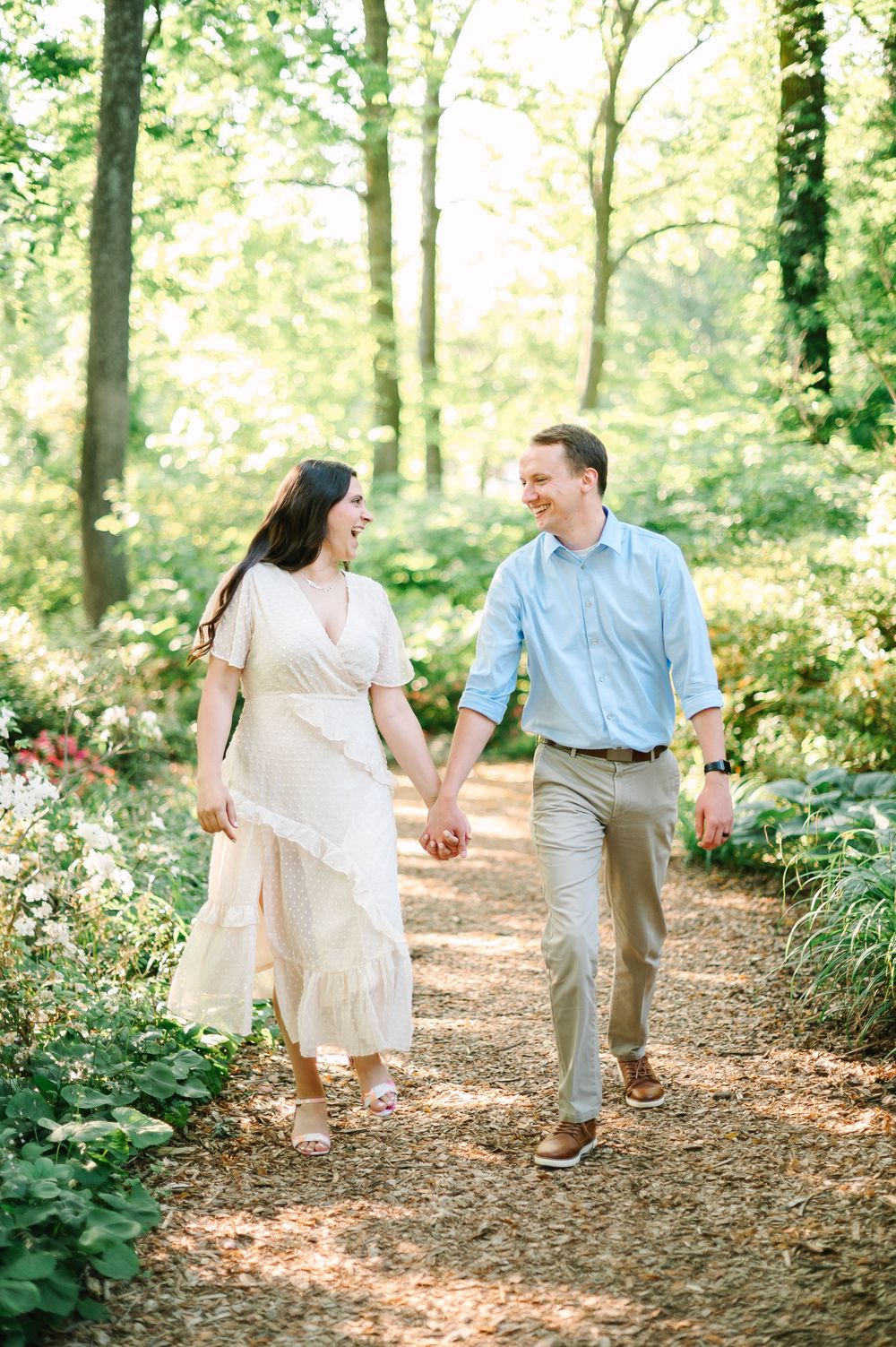 A couple holds hands while walking along a sunlit forest trail.