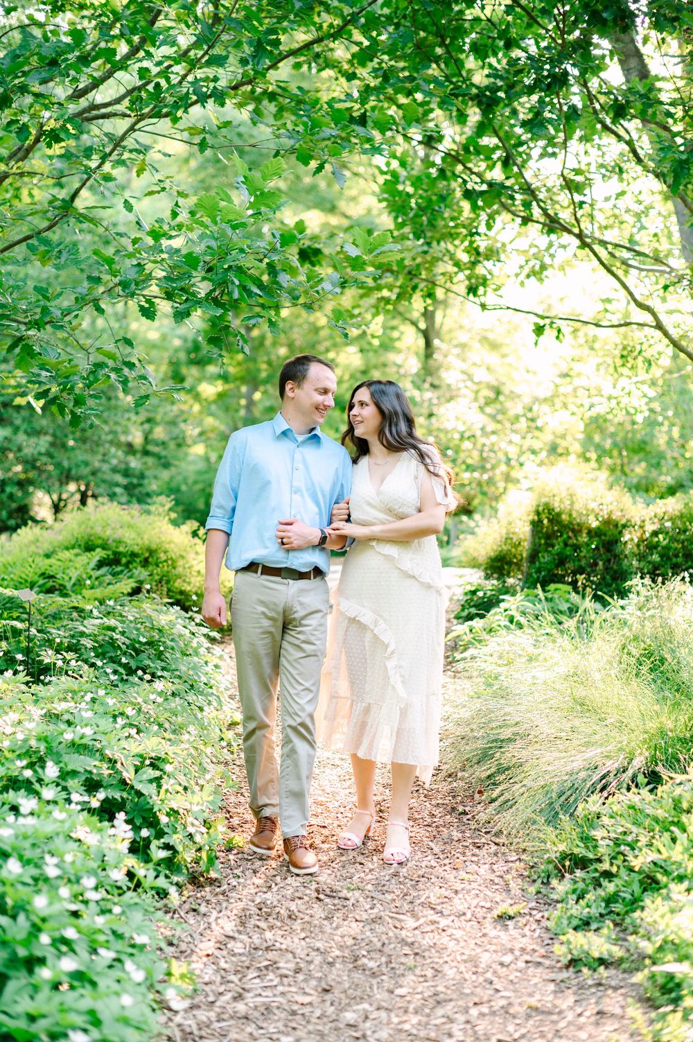 A couple walking together on a leaf-covered path through a lush green garden on a sunny day.
